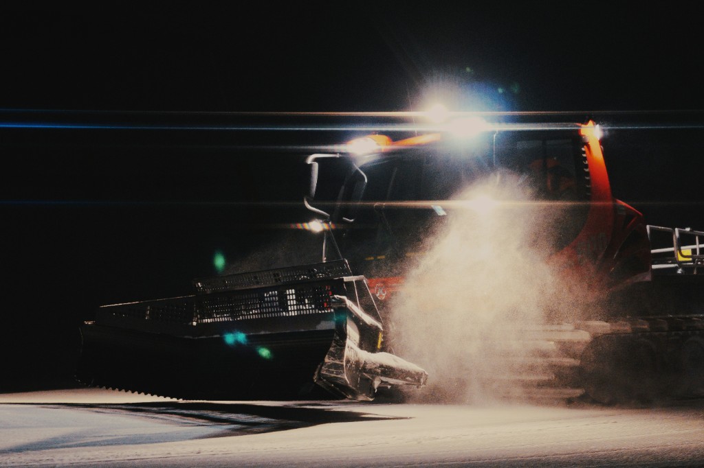 A snow groomer at night, bright work lights and lens flare over blowing snow